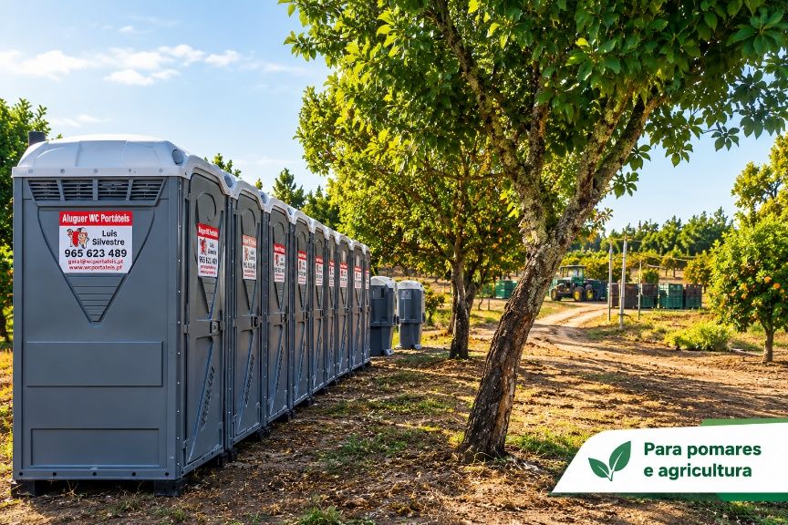 Fila de WC portáteis cinzentos num pomar, com um trator ao fundo