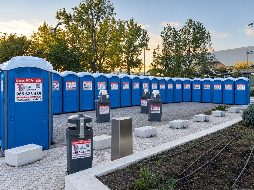 Fila de WC portáteis azuis em curva num recinto de evento, com caixotes do lixo à frente